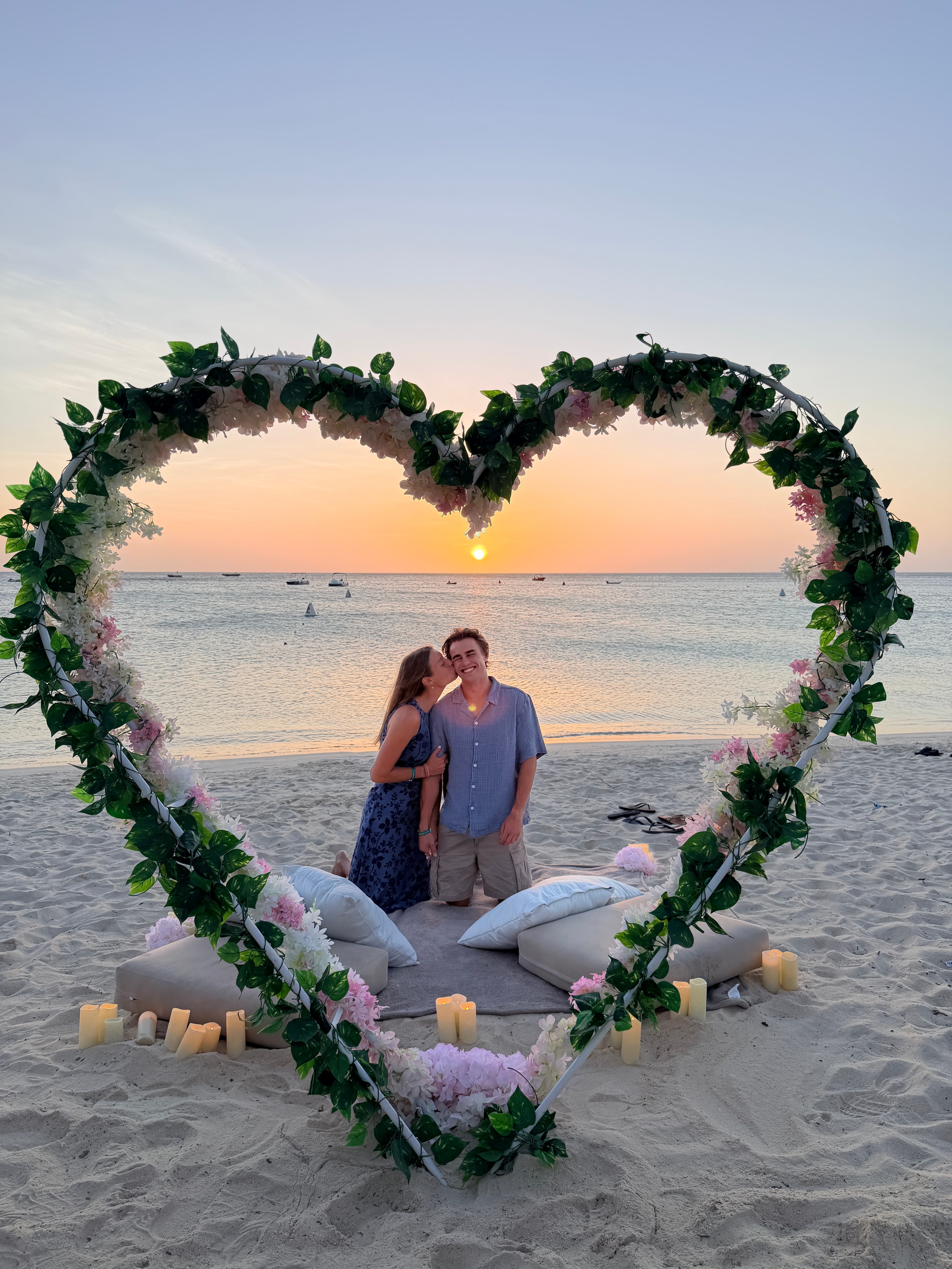 Courtney kissing Carson beneath a flower-covered heart arch on the beach in Aruba at sunset